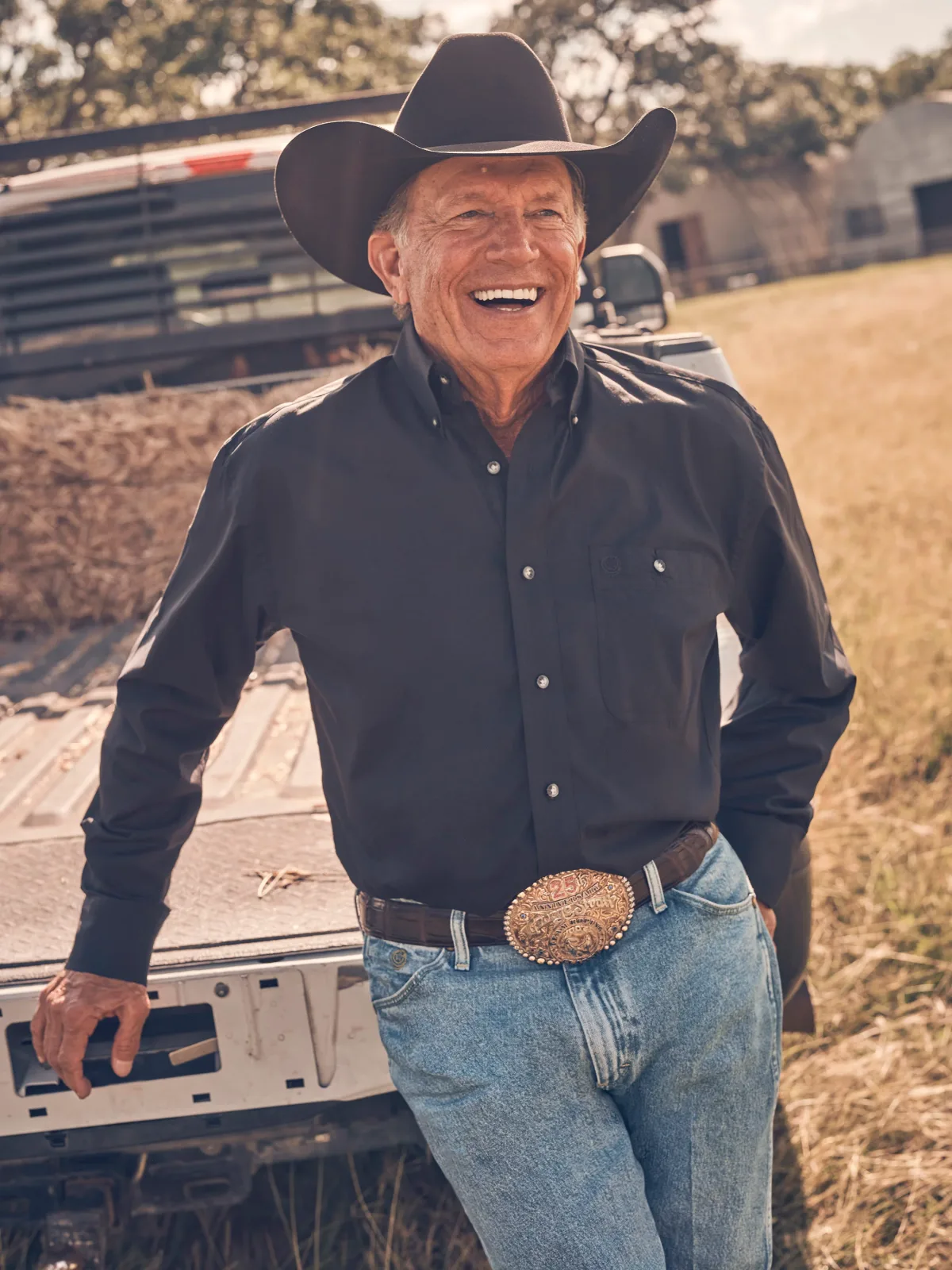 George Strait in a black long sleeve button-down shirt, standing outdoors beside a pickup and hay bales