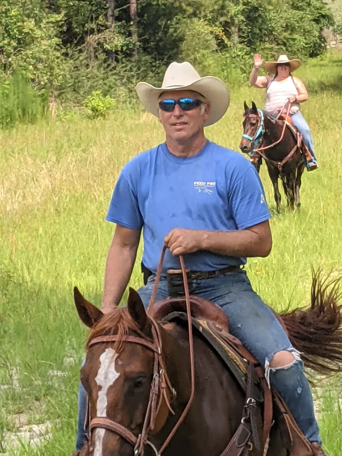 Tony Happel riding a chestnut horse through tall grass in a white cowboy hat, Jennifer Augustine riding behind him.